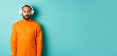 Kulaklıkla müzik dinleyen, sesten etkilenmiş kameraya bakan, turkuaz arka planda duran yetişkin bir adam..