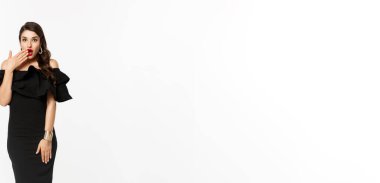 Güzellik ve moda konsepti. Siyah elbiseli, ağzı açık, beyaz arka planda duran ve kameraya bakan güzel ve seksi bir kadının tam boy görüntüsü..