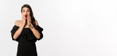 Moda ve güzellik. Siyah elbiseli çekici kadın modelin görüntüsü duyuruya tepki veriyor, kameraya hayret ediyor, beyaz arka planda şaşkın şaşkın duruyor..