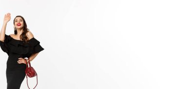 Güzellik ve moda konsepti. Siyah elbiseli ve makyajlı çekici bir kadın. Selam veriyor, gülümsüyor, el sallıyor, beyaz arka plan..