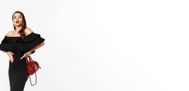 Güzellik ve moda konsepti. Siyah elbiseli, topuklu ayakkabılı, kendine güvenen, kameraya küstah ve beyaz arka planlı zarif bir genç kadın..