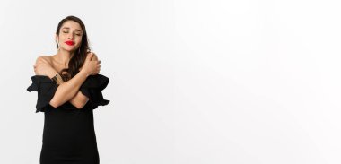 Moda ve güzellik. Siyah elbiseli, şehvetli ve güzel bir kadın, kapalı gözlerle kendine sarılıyor, hayal kuruyor, beyaz arka planda duruyor..