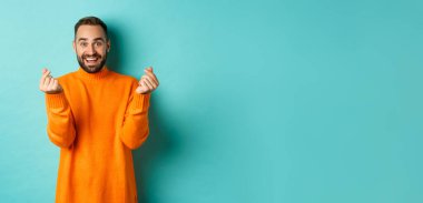 Gülümseyen yakışıklı adam kalp gösteriyor ve mutlu görünüyor, şanslı bir jest, açık mavi arka planda turuncu süveter içinde duruyor..