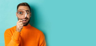 Komik adam büyüteçten bakıyor, bir şeyi arıyor ya da araştırıyor turkuaz arka planda turuncu süveter giyiyor..