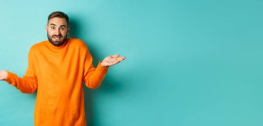 Kafası karışmış, sakallı, omuz silkmiş, bihaber görünen, turkuaz arka planda kararsız duran beyaz bir adam..