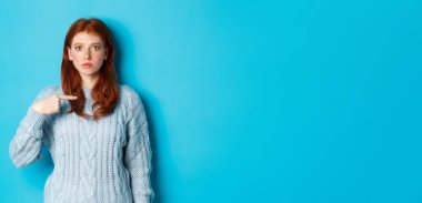 Gergin ve kafası karışık kızıl kız kendini işaret ediyor, mavi arka planda süveter giyiyor..
