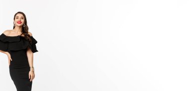 Güzellik ve moda konsepti. Tam boy, güzel esmer kadın, lüks siyah elbiseler ve topuklu ayakkabılar giyiyor, kırmızı dudaklarıyla gülümsüyor, beyaz arka planda dikiliyor..