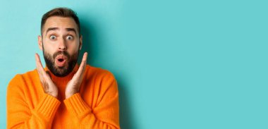 Kameraya bakan, turuncu süveter giyen, turkuaz arka planda duran yakışıklı beyaz adamın yakın çekimi..