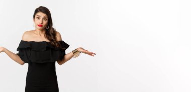 Moda ve güzellik. Siyah elbiseli cahil ve çekici bir kadın. Bilmiyor, omuz silkiyor ve sırıtıyor. Beyaz arka planda kararsız duruyor..