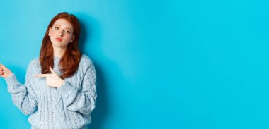 Huysuz genç kız, kızıl saçlı, parmaklarıyla logoyu işaret ediyor, rahatsız ve sıkkın bakıyor, mavi arka planda duruyor..