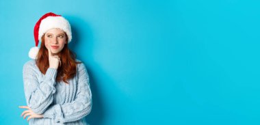 Kış tatili ve Noel arifesi konsepti. Aptal kızıl saçlı kız, Noel Baba şapkası takıyor ve düşünüyor, yeni yıl kutlaması planlıyor, mavi arka planda duruyor..