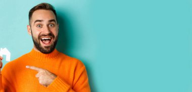 Gayrimenkul. Heyecanlı sakallı bir adam küçük bir kağıt evi işaret ediyor, açık mavi arka planda eğleniyor..