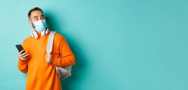 Yüz maskeli genç adam cep telefonu kullanıyor, sırt çantası tutuyor, şaşkın şaşkın bakıyor, açık mavi arka planda duruyor..