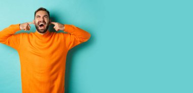 Hüsrana uğramış ve sinirli bir adam üst kattaki komşulara bağırıyor, parmaklarıyla kulaklarını kapatıyor, gürültülü rahatsız edici sesler çıkarıyor, açık mavi arka planda duruyor..