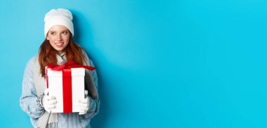 Kış tatili ve Noel satış konsepti. Beyaz bereli ve eldivenli düşünceli kızıl saçlı kadın, elinde bayram hediyesi, sola bakıyor, düşünüyor, mavi arka planda duruyor..