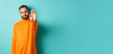 Ciddi somurtkan adam dur işareti yapıyor, hayır diyor, yasaklıyor ve katılmıyor, açık mavi arka planda duruyor..