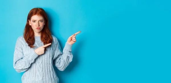 Winter holidays and people concept. Complicated redhead girl staring indecisive, pointing fingers right at logo and frowning, standing perplexed against blue background.