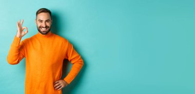 Kendine güveni olan yakışıklı bir adam portakal rengi kazak giymiş iyi bir şey gibi işaret veriyor, onaylıyor ve katılıyor..