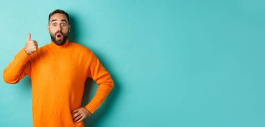 İyi iş çıkardın. Yakışıklı sakallı adam baş parmağını gösterip, iyi işi övüyor, mükemmel bir ürün öneriyor, onaylıyor ve onaylıyor, gülümsüyor, açık mavi arka planda duruyor..