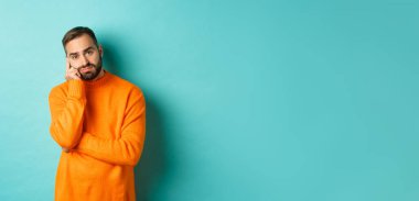 Üzgün ve kasvetli sakallı bir adam, kararsız ve üzgün görünüyor, bir şeyler düşünüyor ve somurtuyor, turkuaz arka planda duruyor..