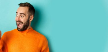 Genç sakallı bir adamın telefon ekranını göstermesi ve hayret verici görünmesi, turuncu süveter giymesi, stüdyo arka planında durması..