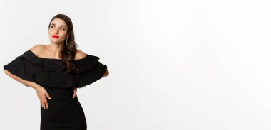 Moda ve güzellik. Siyah elbiseli, makyajlı, kırmızı dudaklı, beyaz arka planda kendine güvenen, beyaz arka planda duran modern bir kadın..