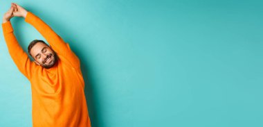 Yakışıklı genç bir adamın güzel bir uykudan sonra ellerini uzatıp gülümsemesi, açık mavi arka planda turuncu kazak içinde durması..