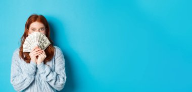 Heyecanlı kızıl saçlı kadın, yüzü parayla kaplı, elinde dolarlarla kameraya bakıyor, mutlu, mavi arka planda dikiliyor..