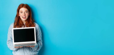 Heyecanlı kızıl saçlı serbest kadın dizüstü bilgisayar ekranı gösteriyor, kameraya şaşkın şaşkın bakıyor, bilgisayarla mavi arka plana bakıyor..