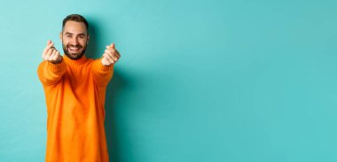 Gülümseyen yakışıklı adam kalp gösteriyor ve mutlu görünüyor, şanslı bir jest, açık mavi arka planda turuncu süveter içinde duruyor..