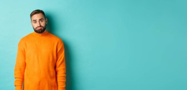 Üzgün adam kameraya bakıyor, somurtuyor ve sıkıntılı hissediyor, açık mavi arka planda duruyor..