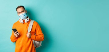 Şüpheli ve hayal kırıklığına uğramış yüz maskesi takan genç adam, sırt çantası ve cep telefonu taşıyor, asık suratlı, açık mavi arka planda duruyor..