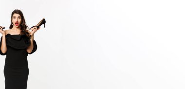 Güzellik ve moda konsepti. Uzun boylu, siyah elbiseli, yüksek topuklu giyen, parti için giyinen, beyaz arka planlı, heyecanlı ve çekici bir kadın..