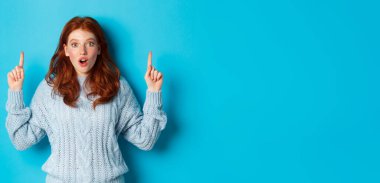 Kış tatili ve insanların konsepti. Şaşkın kızıl saçlı genç kız işaret ediyor, reklam veriyor ve kameraya bakıyor, mavi arka plan..