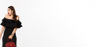 Güzellik ve moda konsepti. Uzun boylu, zarif elbiseli, topuklu ayakkabılı, kafası karışmış, çantasını tutan, ne olduğunu anlamayan, beyaz arka planlı bir kadın..