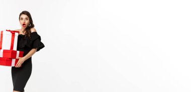 Cazibeli elbiseli, kırmızı dudaklı, elinde Noel hediyeleri olan ve şaşırmış görünen, hediyeler alan, beyaz arka planlı çekici bir kadın model..
