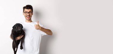 Neşeli yakışıklı bir adam köpeği tutuyor ve tamam işareti gösteriyor, onaylıyor ya da öneriyor. Hipster adam siyah pug taşıyor ve mutlu görünüyor, beyaz arkaplan..