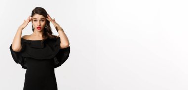 Moda ve güzellik. Siyah elbiseli, kızgın ve yorgun bir kadın. Başa dokunuyor ve yuvarlanıyor. Beyaz arka planda sıkıntılı duruyor..