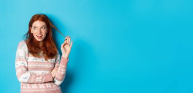 Flört eden kızıl saçlı kız, sola bakıyor, saçlarıyla oynuyor ve düşünüyor, mavi arka planda duruyor..