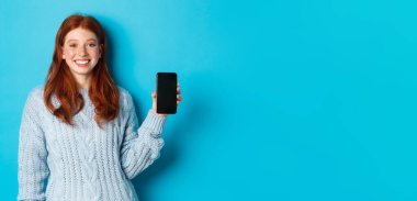 Gülümseyen, kızıl saçlı, akıllı telefon ekranı gösteren, telefonu tutan ve uygulamayı gösteren, mavi arka planda duran bir model..