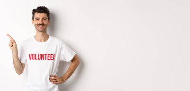 Yakışıklı, gülümseyen, tişörtlü, solu işaret eden, anonsu yapan, beyaz arka planda duran gönüllü..