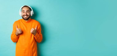 Kulaklıkla müzik dinleyen hayret verici yetişkin bir adam, kameraya bakıyor. Etkilenmiş ve onaylanmış. Tavsiye ediyor, turkuaz arka planda duruyor..