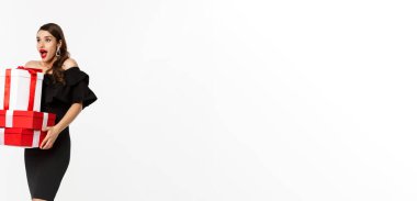 Mutlu Noeller ve yeni yıl tatili konsepti. Siyah elbiseli, heyecanlı ve mutlu bir kadın elinde Noel hediyeleri, logoya şaşırmış bir şekilde bakıyor. beyaz arkaplana karşı sunumlarla ayakta.