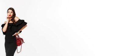 Güzellik ve moda konsepti. Siyah elbiseli güzel ve şık bir kadın. Alışverişi düşünüyor. Sol üst köşeye bakıyor..