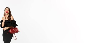 Güzellik ve moda konsepti. Siyah kokteyl elbisesi giymiş, elinde çanta, makyaj yapmış, kameraya gülüyor, beyaz arka planda dikiliyor..