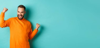 Kazandım. Heyecanlı adam zafer kazanıyor, yumruk tokuşturuyor ve evet diyor, zafer kazanıyor ve tatmin olmuş görünüyor, hedefe ulaşıyor, açık mavi arka planda duruyor..