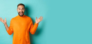 Heyecanlı adam reklam teklifine tepki veriyor, ellerini yana açıyor ve seviniyor, turkuaz arka planda turkuaz süveter giyiyor..