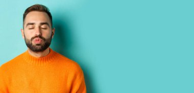 Süveter giyen, dudaklarını büzen, gözlerini kapayan ve öpücüğü bekleyen, açık mavi arka planda duran genç adamın yakın çekimi..