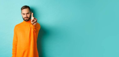 Ciddi sakallı adam kaşlarını çatıyor, hareket yasağı koymak için parmağını sallıyor, bir şeyi yasaklıyor, hayal kırıklığını dile getiriyor, açık mavi arka planda seninle aynı fikirde değil..