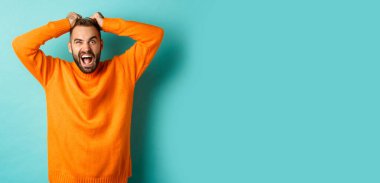 Sinirli adam bağırıyor, saçlarını yoluyor, bağırıyor, sinirleniyor, sinirleniyor ve açık mavi arka planda dikiliyor..